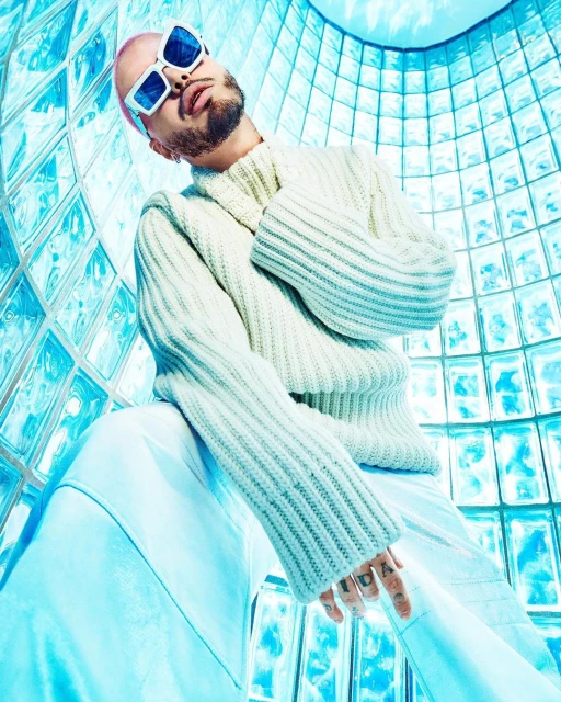 Man with buzzcut and sunglasses wearing a cream-colored sweater poses in front of translucent blue glass brick background.