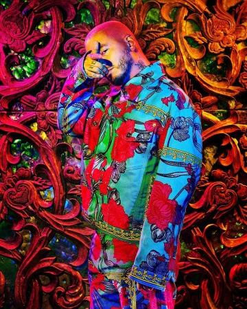 Man in a colourful patterned jacket stands in front of carved wooden panels. The image features detailed patterns and vibrant colours.