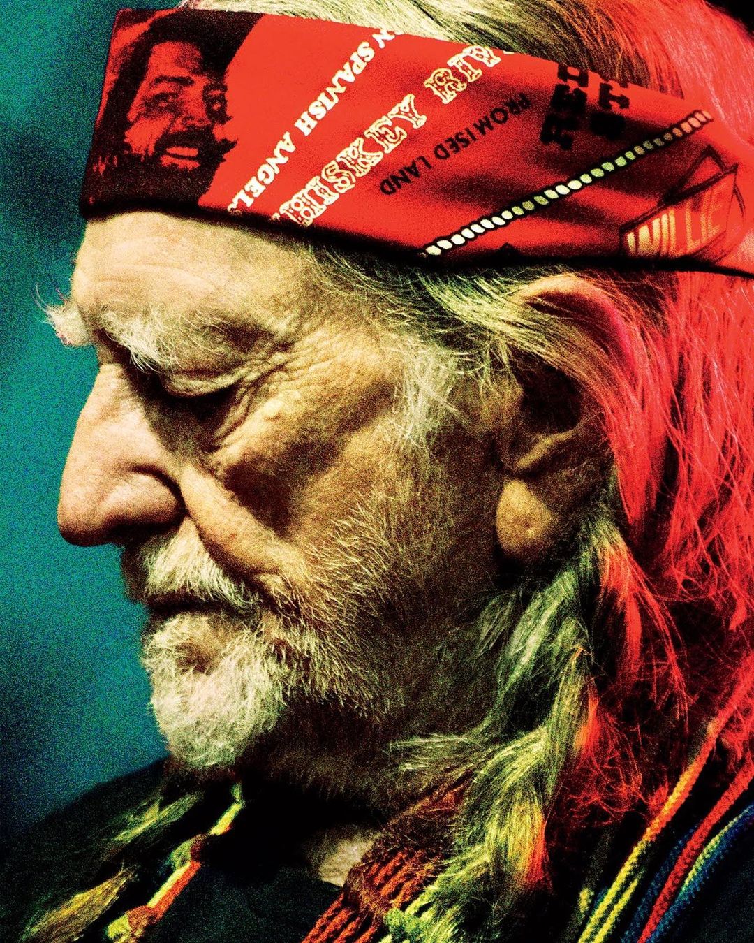 Close-up profile portrait of an elderly man with a red bandana, long white hair and beard, and a multicolored scarf.