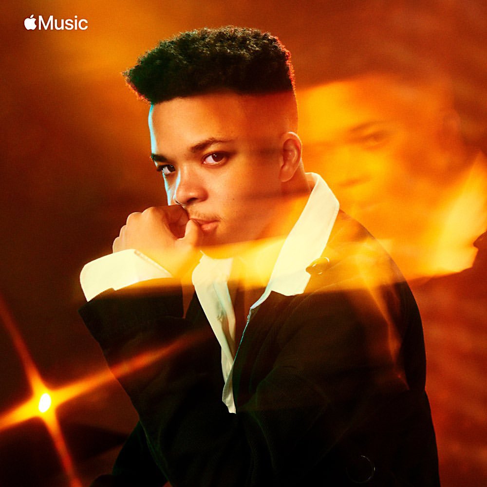 Portrait of a young Black man in a suit jacket, with a hand near his mouth, against an orange, softly-lit background. Apple Music logo visible.
