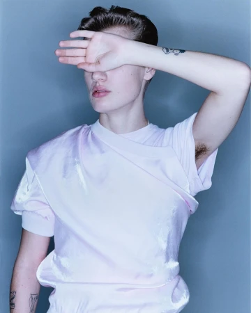 Portrait of a person with short brown hair, wearing a pink shirt and tattoos, covering their face with their hand in a photo studio.