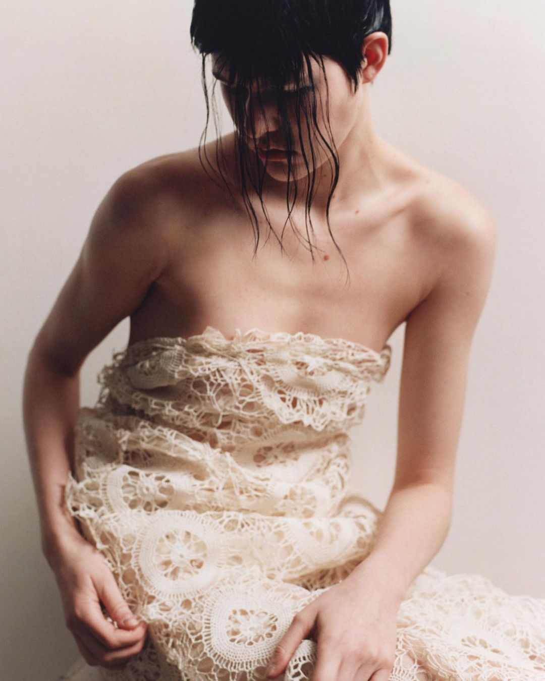 Portrait of a young adult woman with wet dark hair, wearing a lace garment, set against a neutral background, fashion editorial.