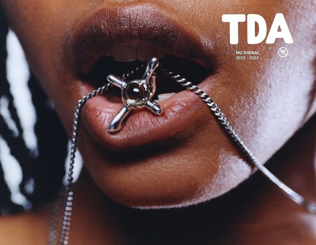 Close-up of a woman with brown skin and dark hair, wearing a metal necklace with a cross-shaped pendant in her mouth.