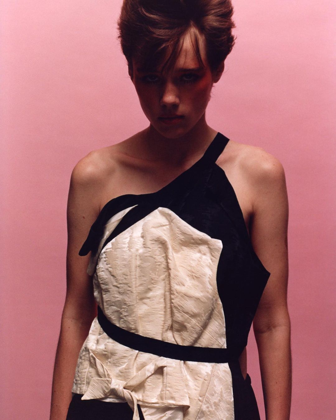 Fashionable adult woman in a black and cream dress poses against a pink background in a studio portrait shot indoors.