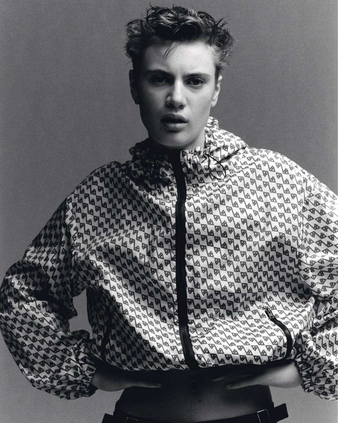 Portrait of a young adult female with short hair, wearing a patterned jacket, set against a neutral gray background in a studio setting.