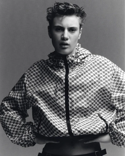 Portrait of a young adult female with short hair, wearing a patterned jacket, set against a neutral gray background in a studio setting.