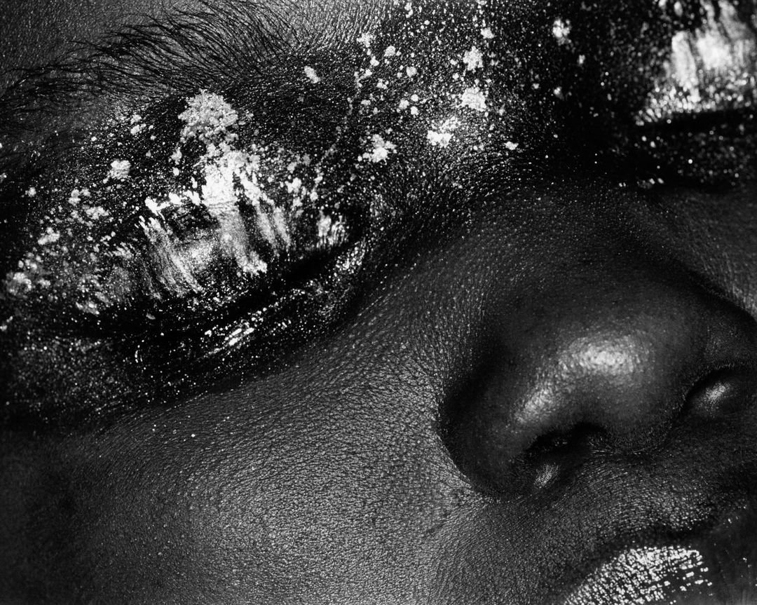 Close-up black and white portrait of a woman's face with eye closed, wearing makeup with white glitter on her eyebrow and eyelid.