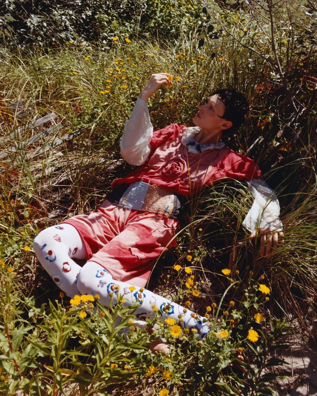 Person in a red outfit with patterned tights lying in tall grass with yellow wildflowers, bathed in bright sunlight in an outdoor setting.