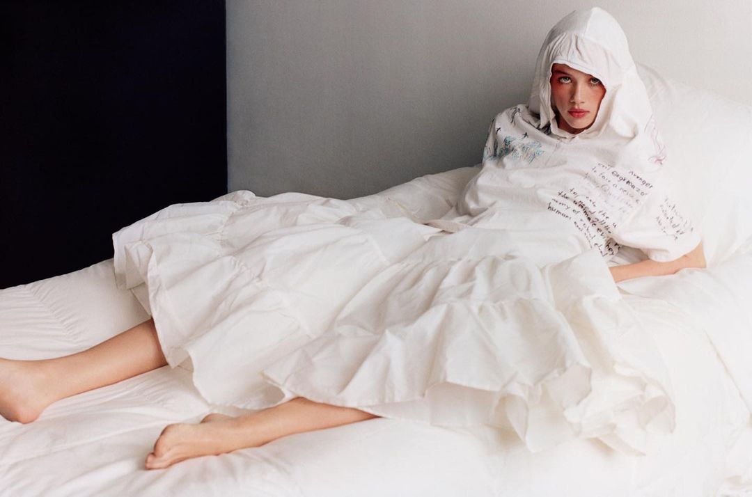 Adult female lies on a white bed in a hooded top. She has her legs extended and looks directly at the viewer in this indoor shot.