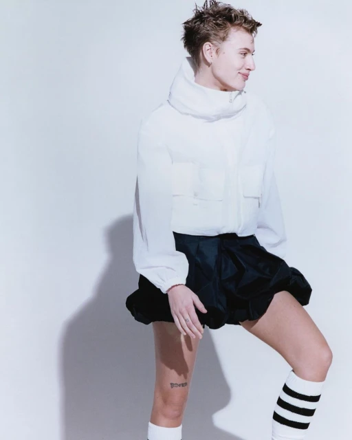 Adult posing in a white puffer jacket, black skirt, and striped socks against a white background in a studio environment. Fashion editorial.