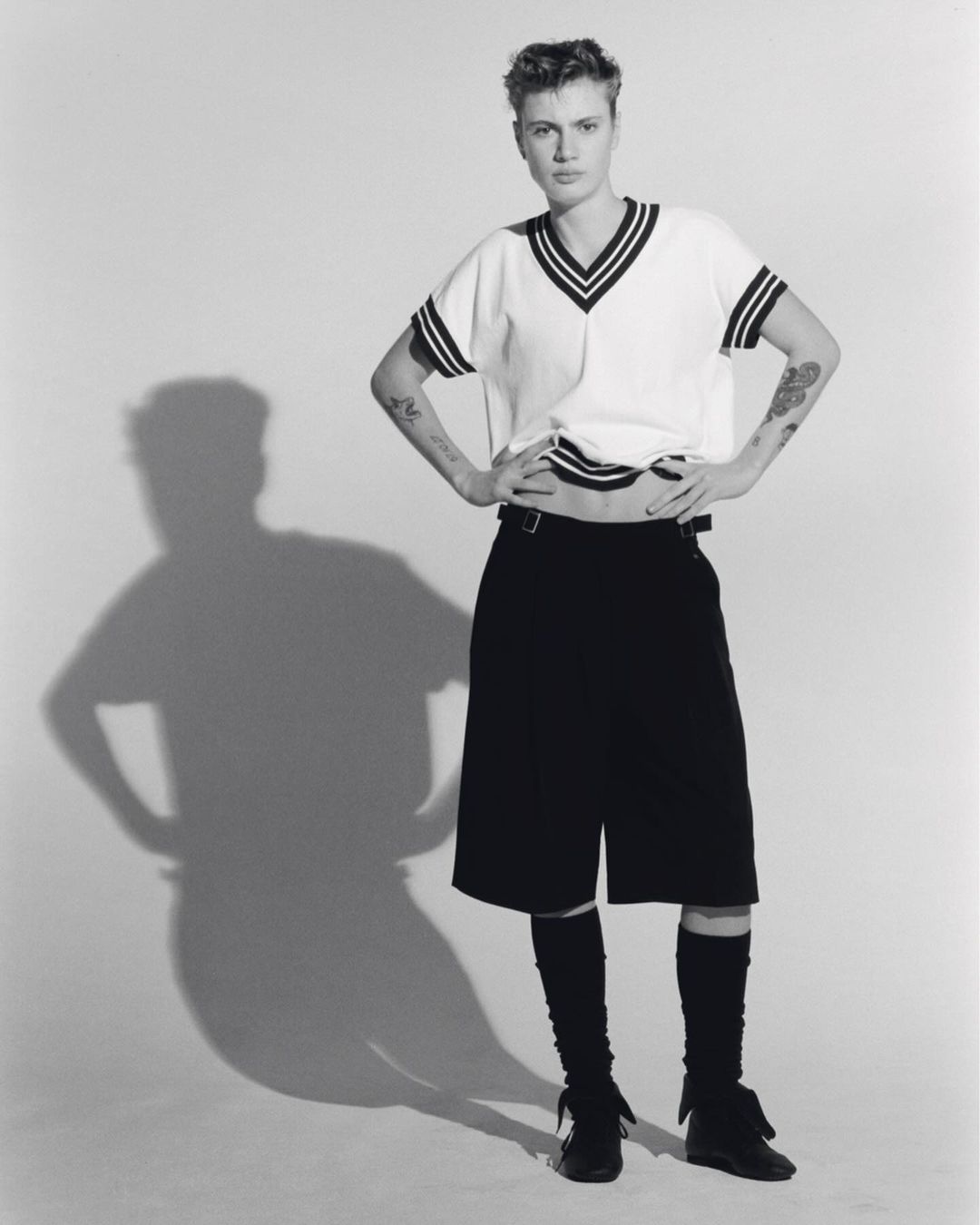 Black-and-white studio shot of a young adult woman in a short-sleeved sweater, shorts, and leg warmers, posing with hands on hips.