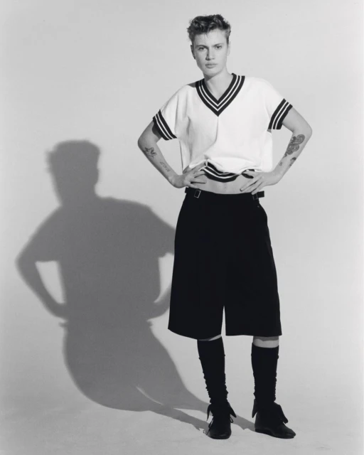 Black-and-white studio shot of a young adult woman in a short-sleeved sweater, shorts, and leg warmers, posing with hands on hips.