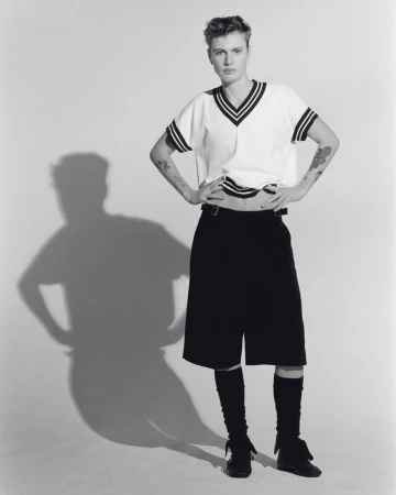 Black-and-white studio shot of a young adult woman in a short-sleeved sweater, shorts, and leg warmers, posing with hands on hips.