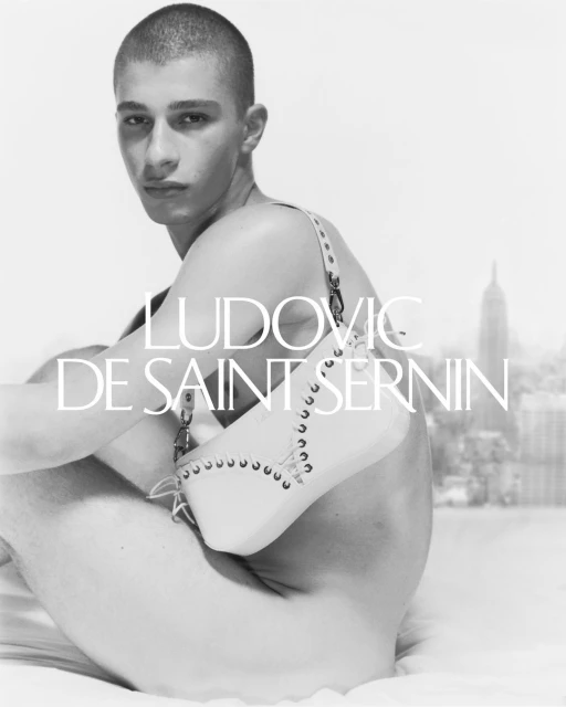 Black and white studio shot of a young adult male posing with a studded leather purse in front of a blurred city skyline.