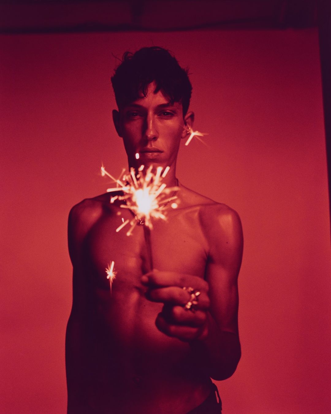 Portrait of a slender, bare-chested young adult holding a lit sparkler against a stark red backdrop, emphasizing contrast and mood.
