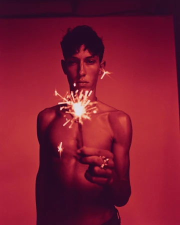 Portrait of a slender, bare-chested young adult holding a lit sparkler against a stark red backdrop, emphasizing contrast and mood.