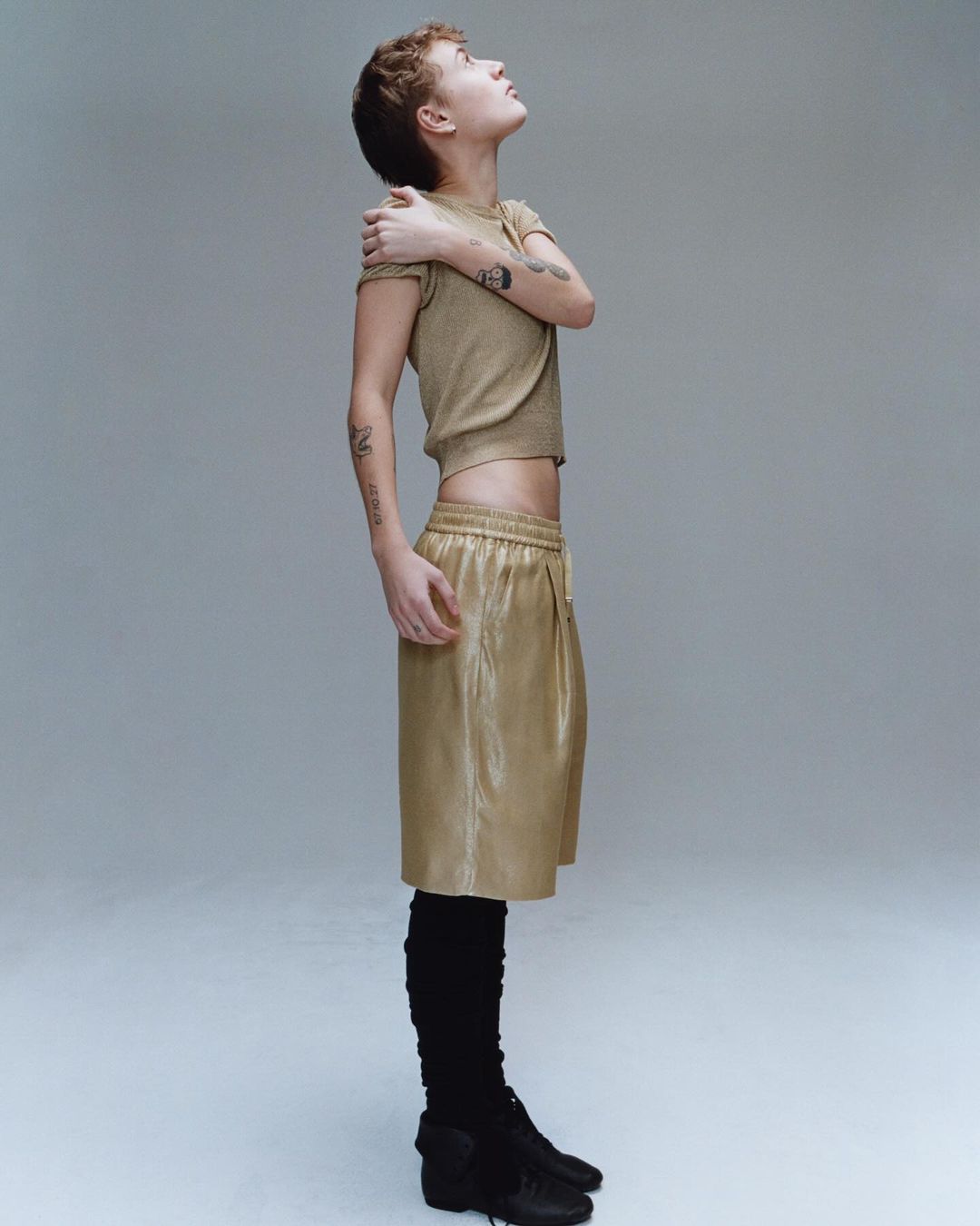 Side profile of a young adult woman with short hair in a crop top and shorts, looking up in a studio with a neutral background.