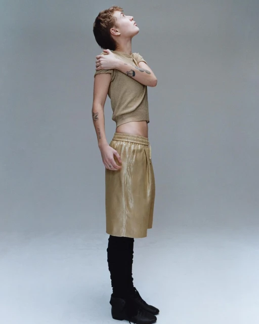 Side profile of a young adult woman with short hair in a crop top and shorts, looking up in a studio with a neutral background.