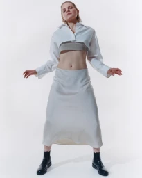 Model wearing a cropped white shirt, grey top, long skirt, and black boots against a white background by Parker Woods