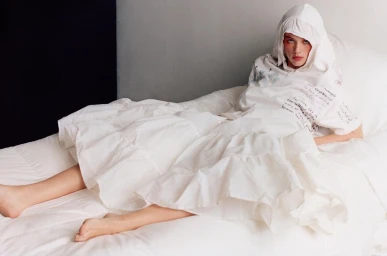 Model lying on a white bed wearing a white hooded dress with text print and layered ruffles, photographed by Parker Woods.