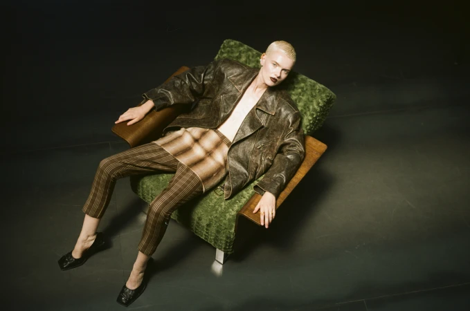 A blonde model with dark lipstick wearing a leather jacket and striped trousers seated in a green armchair