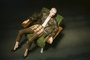 A blonde model with dark lipstick wearing a leather jacket and striped trousers seated in a green armchair