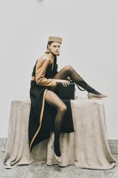 Model wearing pillbox hat and black coat sitting on a cloth-covered table holding a black box bag