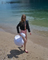 Model in dark sunglasses carrying a white swan prop along a sandy beach beside clear water