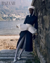 Model in dark coat and white sculptural peplum leaning against a stone wall by a lake, photographed by Albina Abdullina.