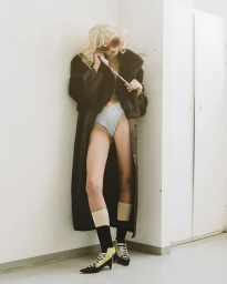 Model wearing a dark fur coat, sunglasses, and high heels standing against a white wall while on a telephone.