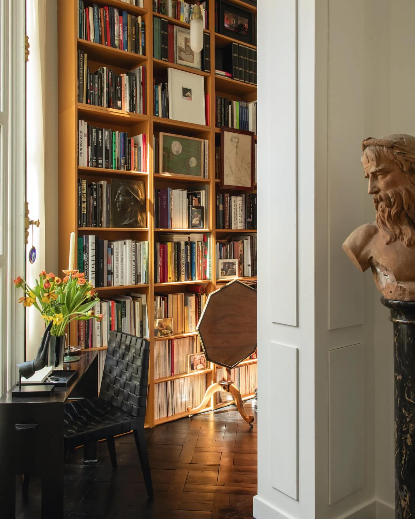 Home library featuring a large bookcase filled with books, a black writing desk with flowers, and a bust sculpture in a well-lit room.