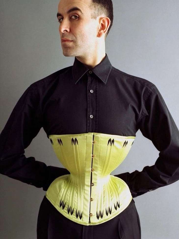 Adult male poses indoors wearing a light-yellow corset over a black button-down shirt and black pants against a grey backdrop.