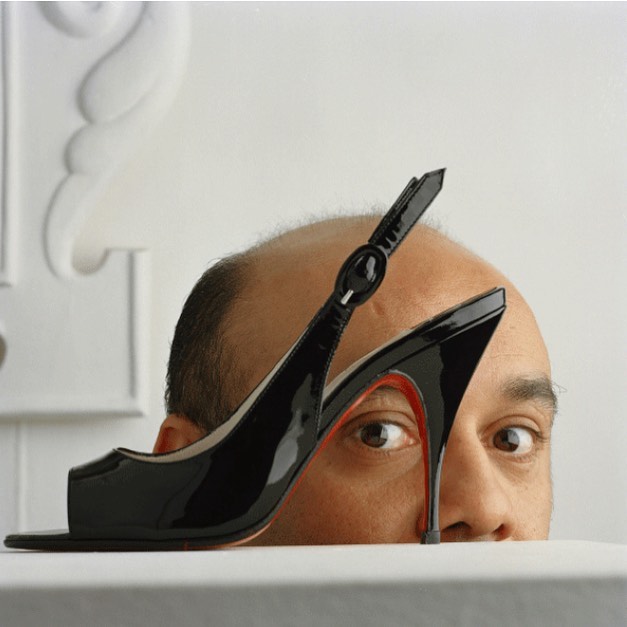 A glossy black slingback heel rests in front of a fair-skinned bald man. The heel has a red sole. White background with subtle mouldings.