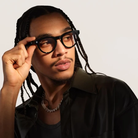 Close-up portrait of a man wearing black frame glasses and a dark button-down shirt against a plain light background.