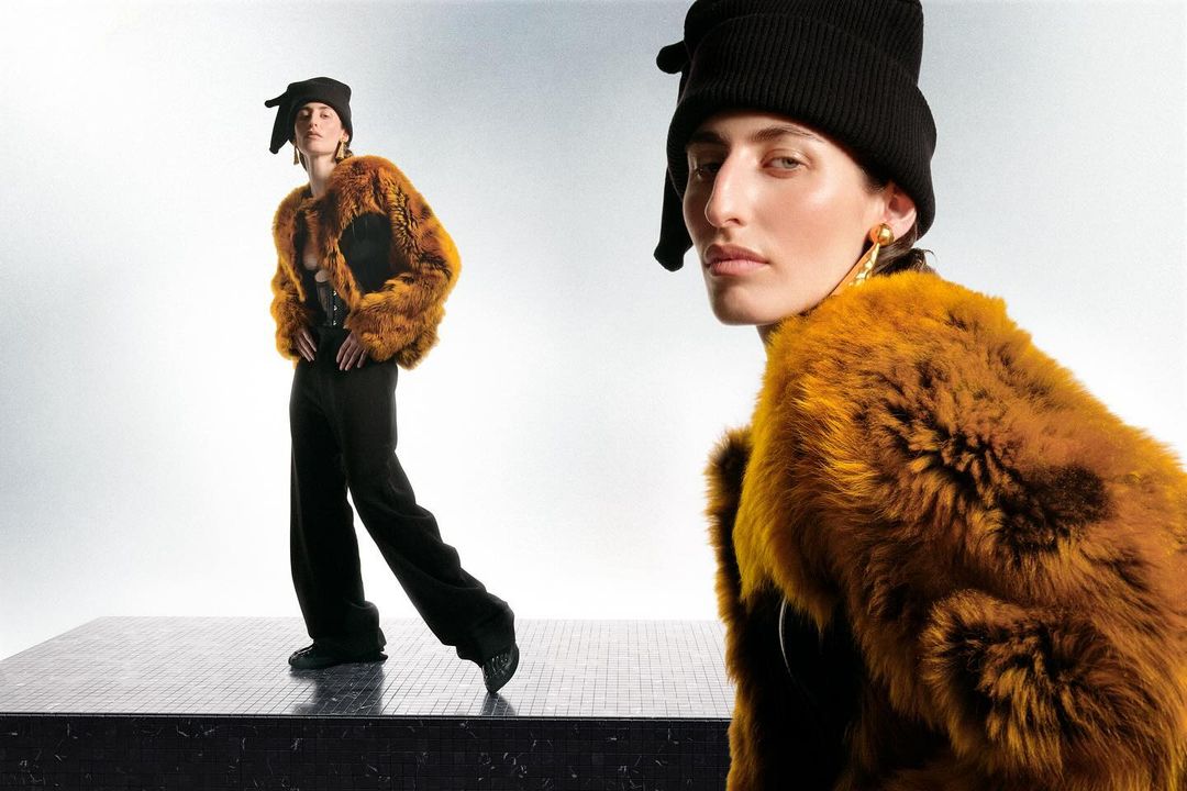 Two adult female fashion models pose in fur coats, black knit caps, and pants on a dark tile platform in a light grey photo studio.