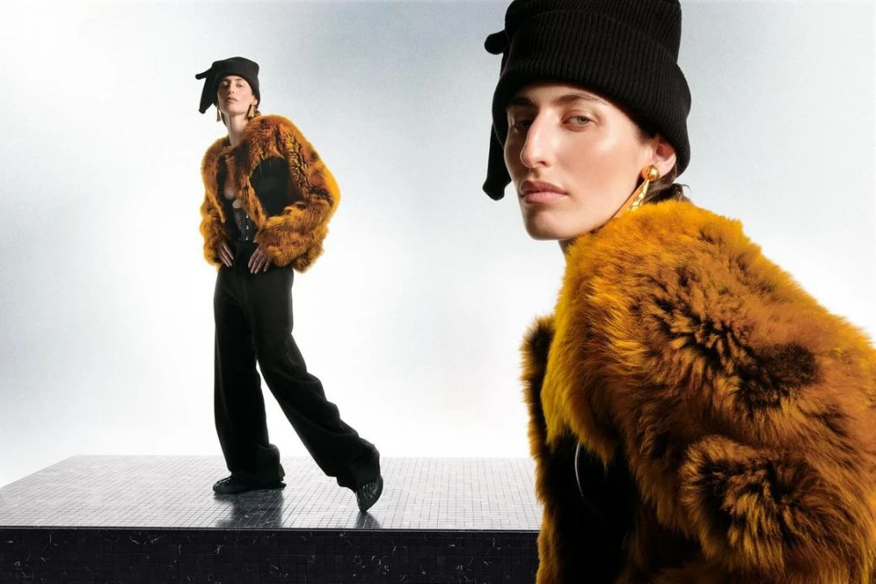 Two adult female fashion models pose in fur coats, black knit caps, and pants on a dark tile platform in a light grey photo studio.