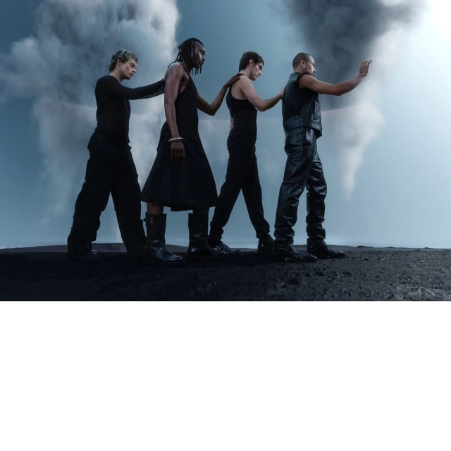 Four adult male fashion models pose together in a line outdoors against a cloudy sky wearing dark, monochromatic clothing and boots.