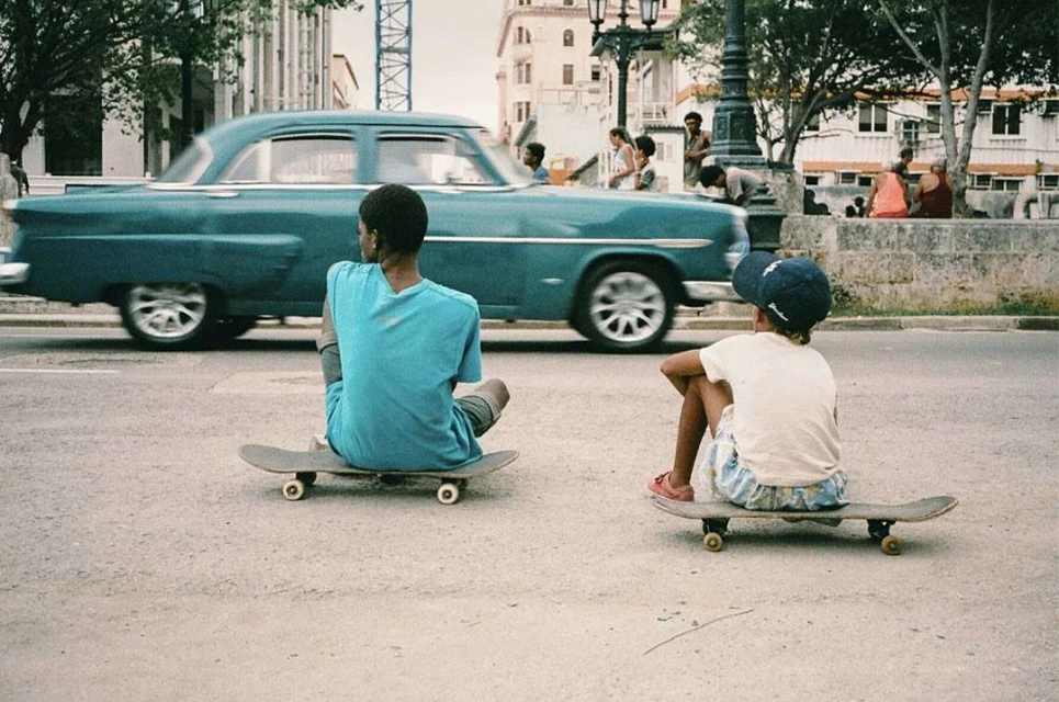 Two kids on skateboards sit in a city street; a vintage car passes in the background with other people on the sidewalk.