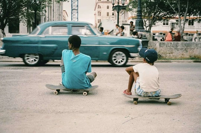Two kids on skateboards sit in a city street; a vintage car passes in the background with other people on the sidewalk.