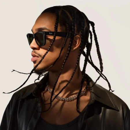 Profile portrait of a black man wearing black sunglasses and a dark leather shirt against a white seamless backdrop.