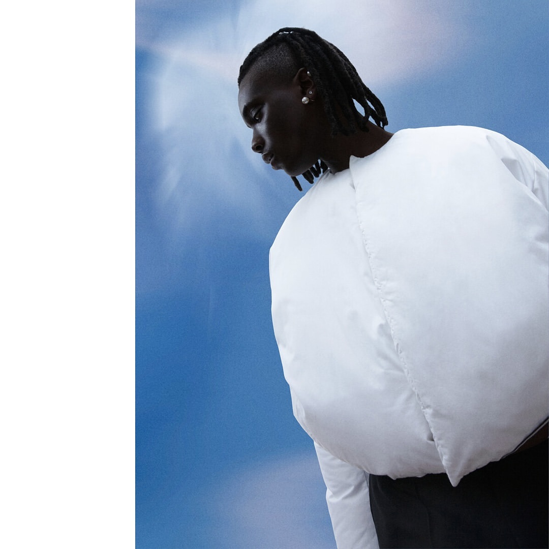 Portrait of a Black male model in a white puffer jacket against a blue cloudy sky. Fashion shot with natural light and a low angle.
