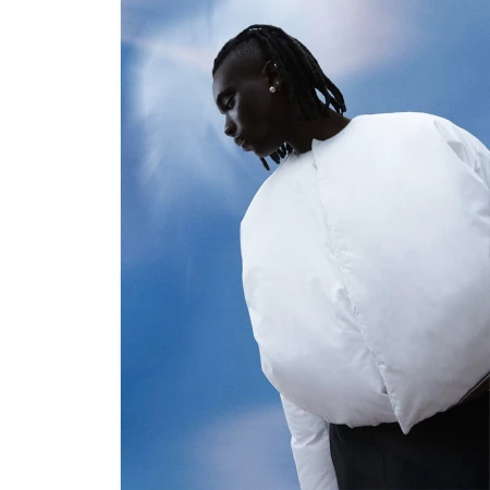 Portrait of a Black man with dreadlocks wearing a white puffy jacket against a cloudy blue sky background.