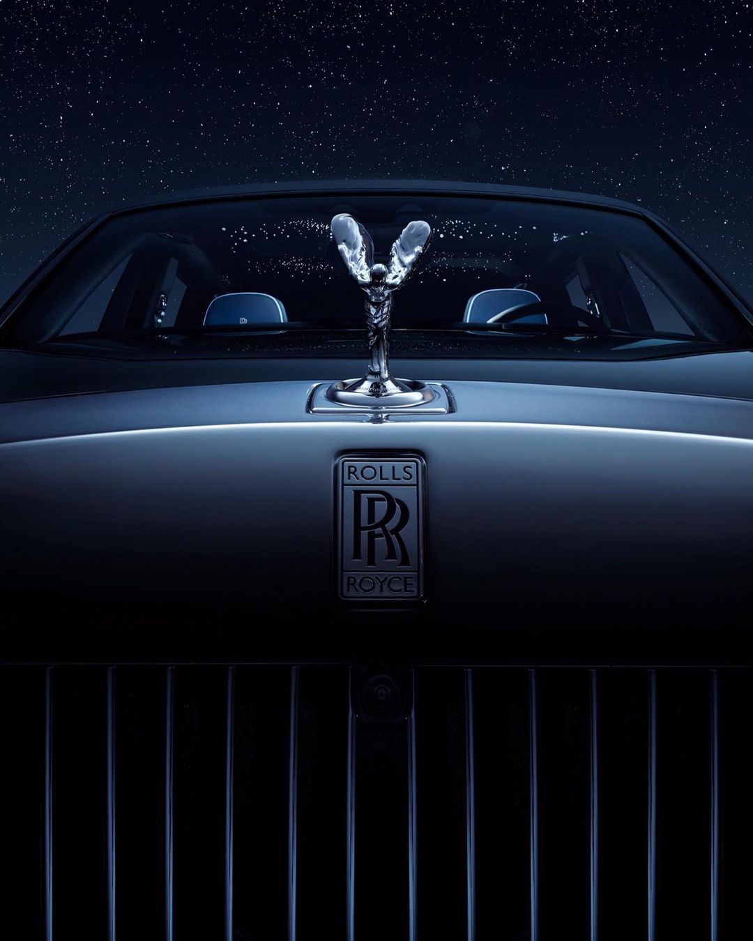 Silver Rolls Royce front view with the Spirit of Ecstasy hood ornament under a night sky filled with stars. Automotive elegance.