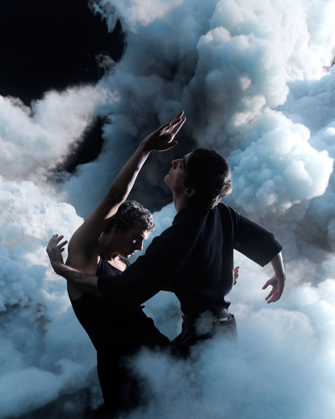 Two athletic adult humans, possibly dancers, embrace in a fog-filled dark space with arms raised in a modern dance-inspired performance.