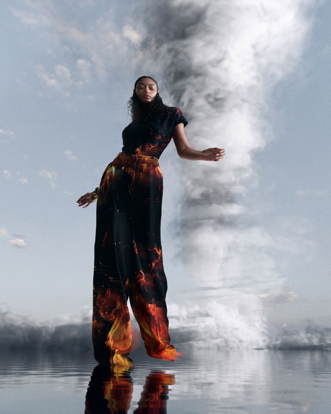 A woman poses in a black and orange fire-printed outfit, standing on reflective water under a blue sky with a swirling smoke cloud.