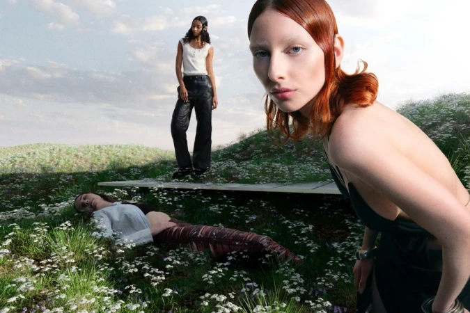 Three fashion models pose in an outdoor setting with a grassy hill landscape; one model is lying down, another is standing on a bench.
