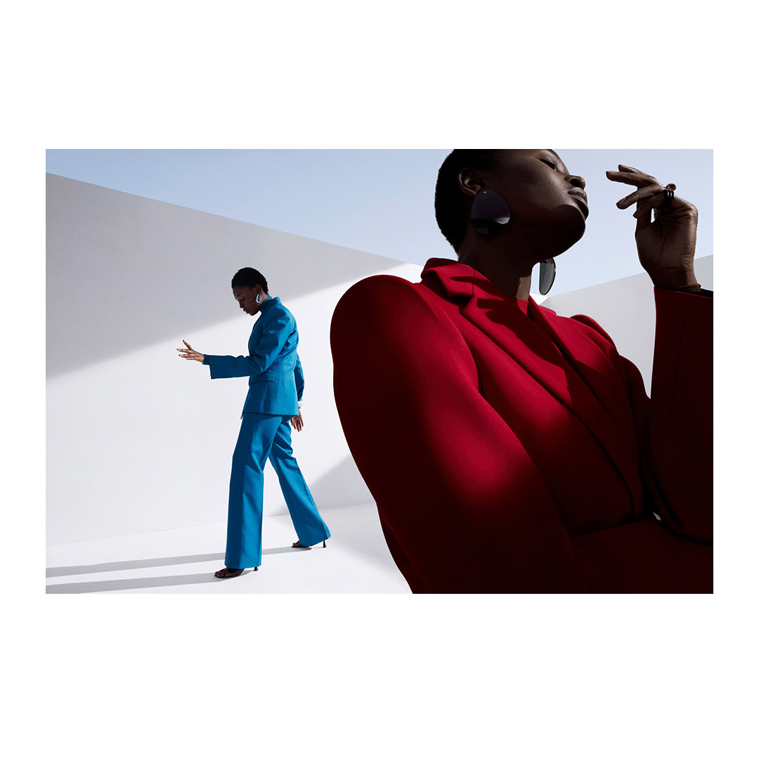 Two African American women in a minimalist studio setting, one in a red suit facing the camera and the other in a blue suit.
