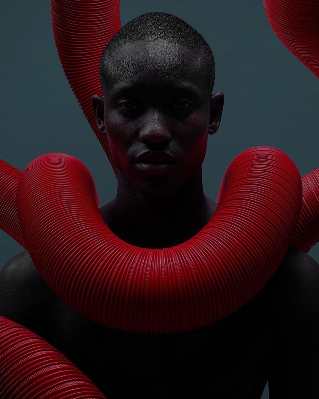 Close-up studio shot of an adult male with dark skin surrounded by red ribbed tubes. The subject gazes directly forward in a portrait orientation.