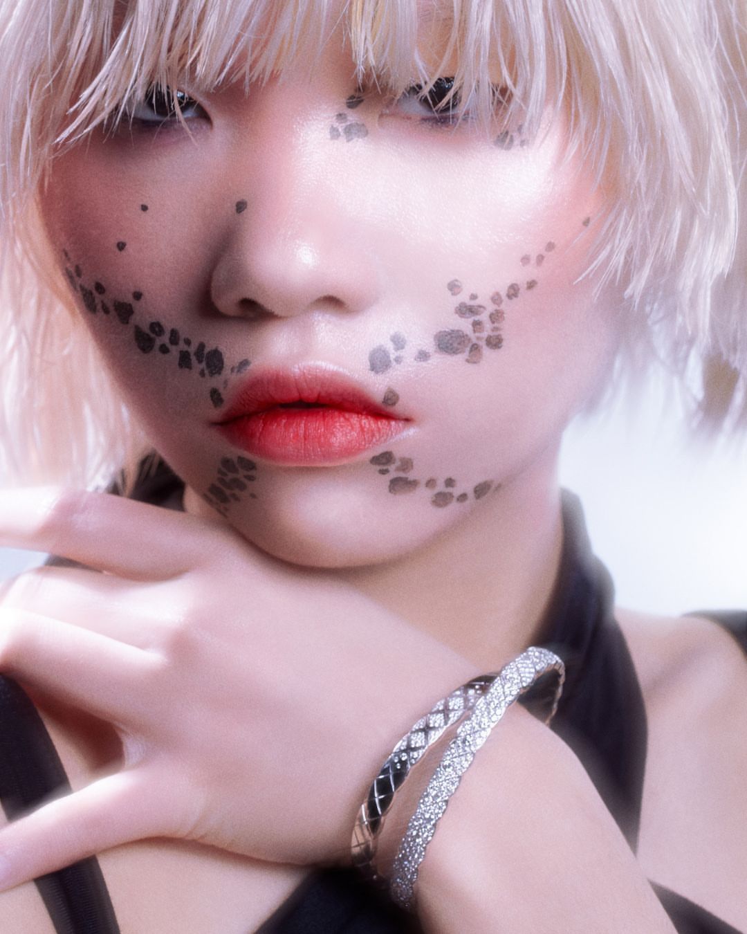 Close-up of a fair-skinned person with blonde hair, red lips, and black dot face makeup wearing a black top and sparkling bracelets.
