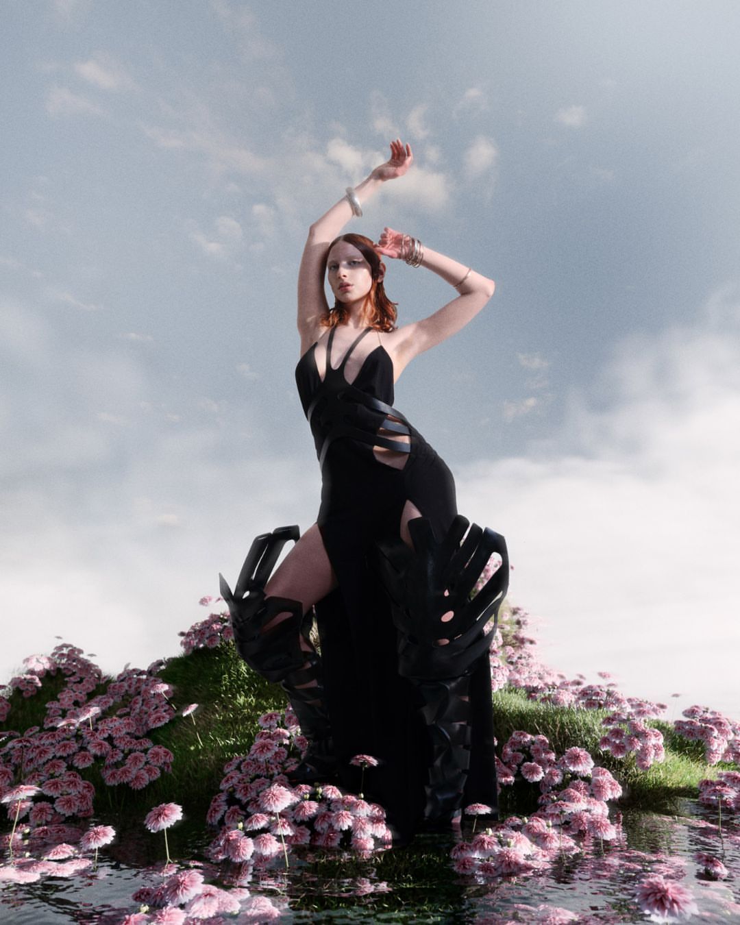 Woman in a black dress standing in a field of pink flowers under a cloudy sky, wearing decorative arm cuffs and boots.